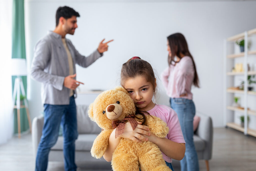 Family conflicts. Little girl suffering from parents quarrels, cuddling her teddy bear, feeling lonely and depressed. Stressed kid looking at camera, selective focus on child