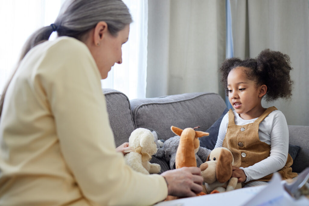 Psychologist, kid and teddy bear in office for therapy, ADHD assessment and notes on sofa. Mature therapist, girl and stuffed animals in practice for evaluation, sensory development and psychology.