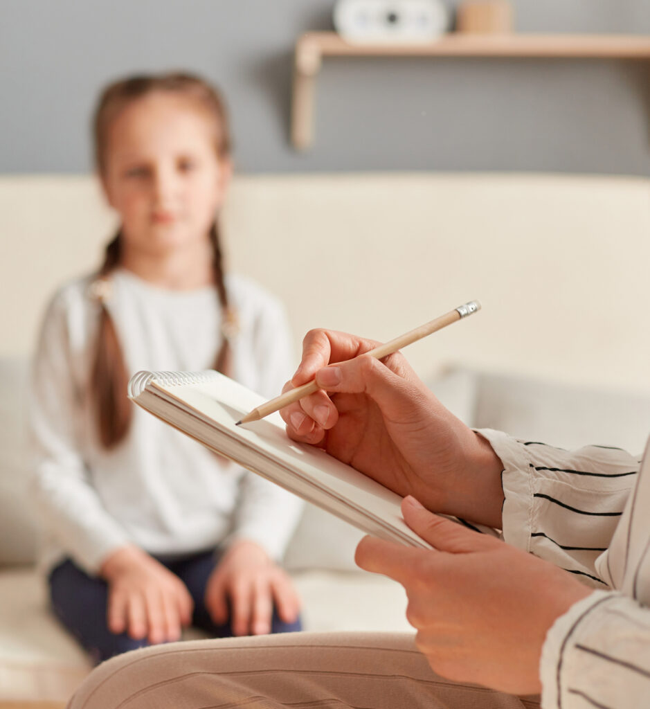 Psychologist taking notes while conducting session with female child, writing down important points about her emotional and mental condition, listening about little girl problems.